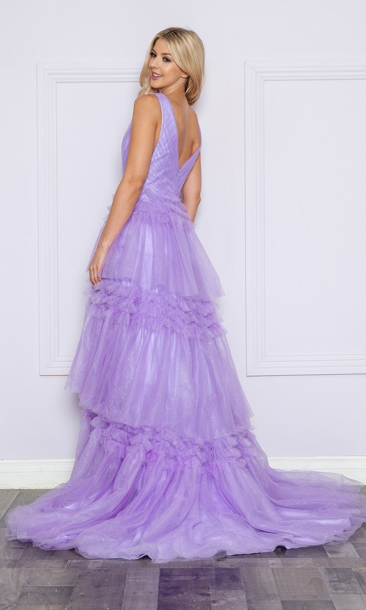 A woman stands indoors wearing the Poly USA 9406 Ruffled Long V-Neck Prom Ball Gown, a floor-length A-line dress with tulle tiers and a fitted bodice in lavender, glancing over her shoulder in this elegant evening look.